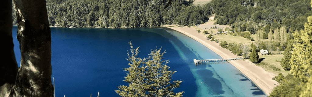 Vista panorámica desde un mirador elevado del Lago Espejo en Villa La Angostura, mostrando una playa de arena, un muelle largo, aguas de azul profundo a turquesa y montañas boscosas bajo un cielo despejado