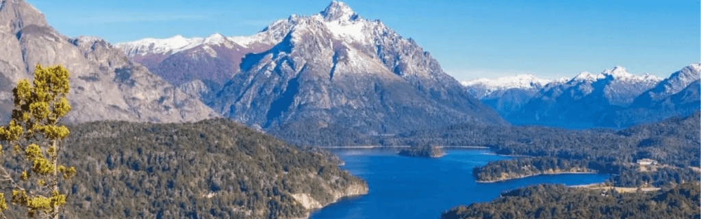 Vista panorámica desde un mirador con baranda de hierro hacia el Lago Nahuel Huapi, rodeado de bosques densos y montañas con cumbres nevadas bajo un cielo azul despejado en Bariloche.