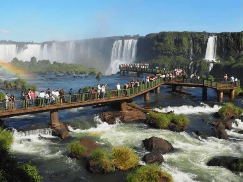 Turistas caminando por las pasarelas metálicas sobre el río Iguazú, con grandes saltos de agua al fondo y un arcoíris formándose en la bruma.