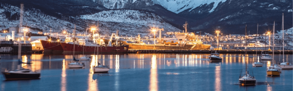 Vista nocturna del puerto de Ushuaia con barcos iluminados en el agua mansa y las imponentes montañas de la Cordillera de los Andes cubiertas de nieve al fondo bajo un cielo azul crepuscular