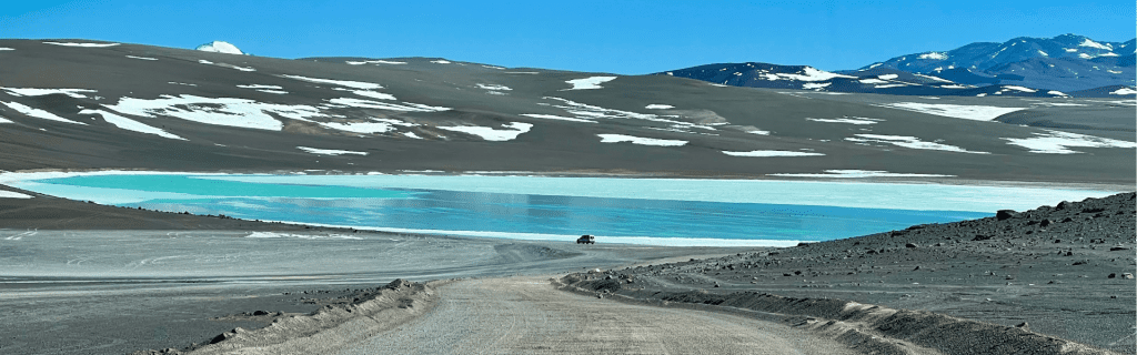 Camino de ripio serpenteando hacia una laguna de color esmeralda en medio de un paisaje lunar de montañas con restos de nieve en Catamarca.