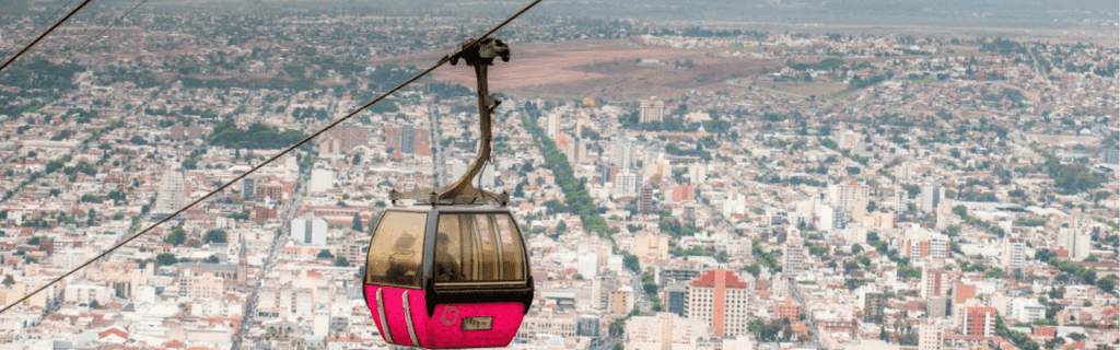 Una góndola roja y gris del teleférico suspendida por cables sobre la ciudad de Salta, con una vista panorámica de las casas y montañas al fondo bajo un cielo cálido al atardecer.