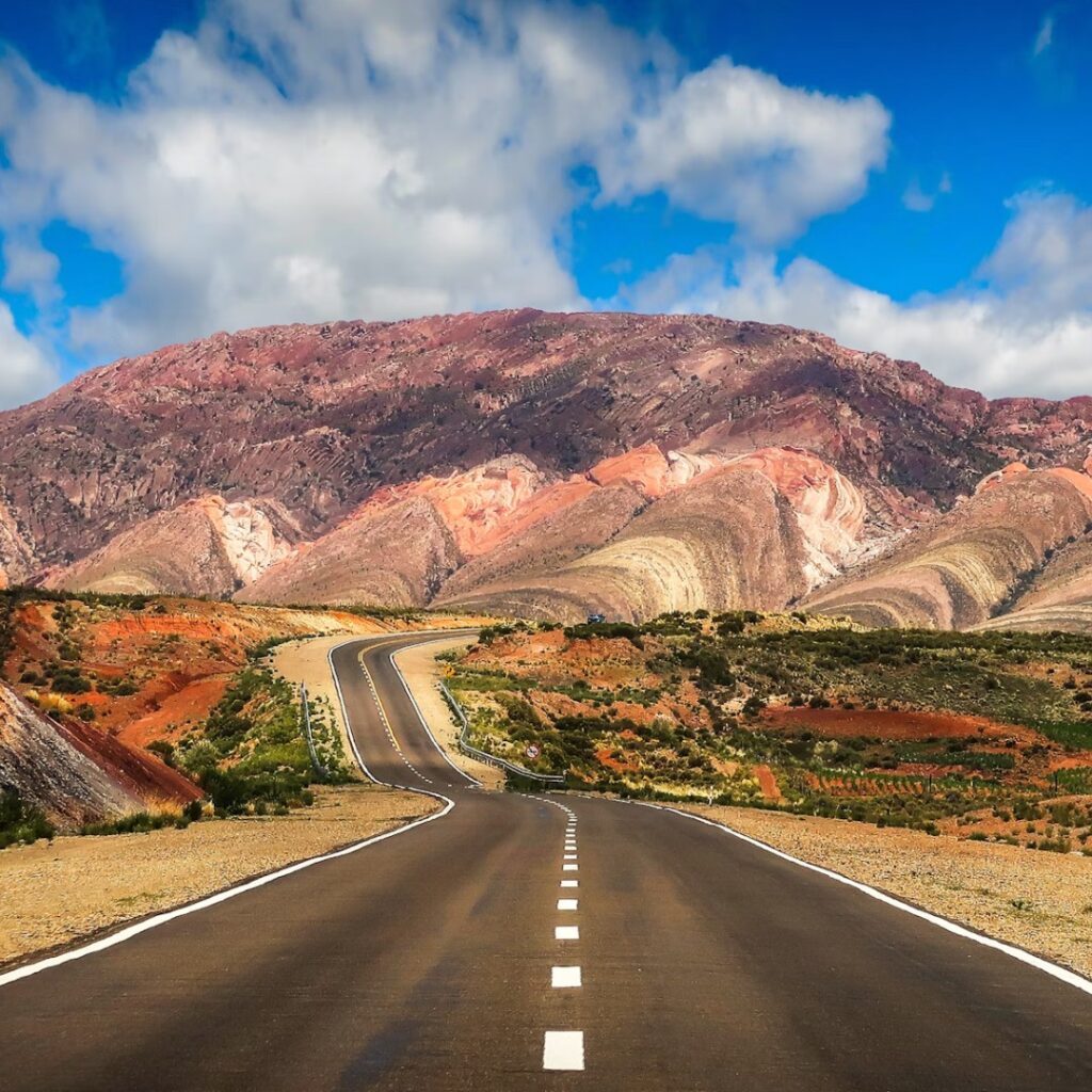 Carretera asfaltada y sinuosa que atraviesa un paisaje de montañas áridas con estratos geológicos de colores rojizos y amarillos bajo un cielo azul con nubes blancas en el Norte Argentino.